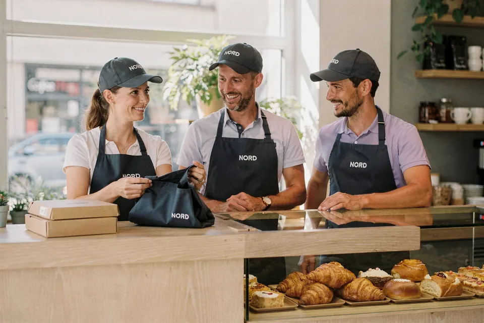 Drei Mitarbeitende einer Franchise-Filiale in einheitlichen Schürzen öffnen ein Paket mit neuer Arbeitskleidung am Tresen.