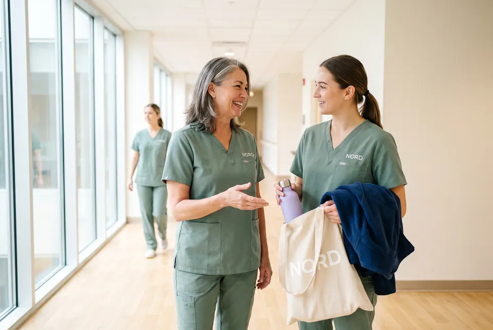 Eine Pflegekraft begrüßt eine neue Kollegin im Klinikflur, beide tragen grüne Kasacks mit Logo, die neue Kollegin hält eine Willkommenstasche.