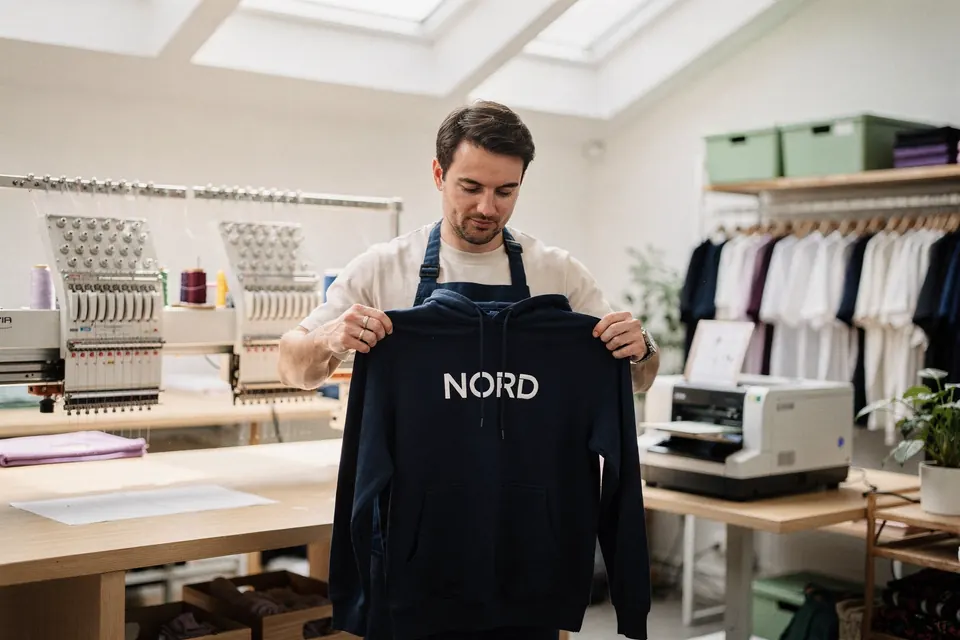 Eine Textilveredlerin prüft in der Werkstatt einen frisch bestickten Hoodie, im Hintergrund Stickmaschine und Lasergravur.