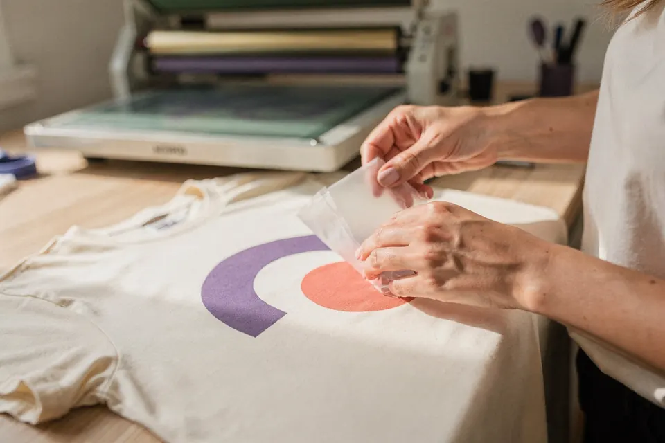 Nach dem Heißpressen zieht eine Hand die Trägerfolie vom T-Shirt und legt den farbigen DTF-Druck frei.