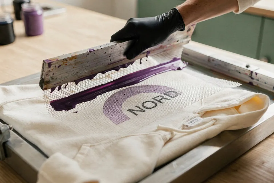 Beim Siebdruck zieht eine Hand den Rakel mit violetter Farbe über das Sieb, darunter liegt ein Hoodie.